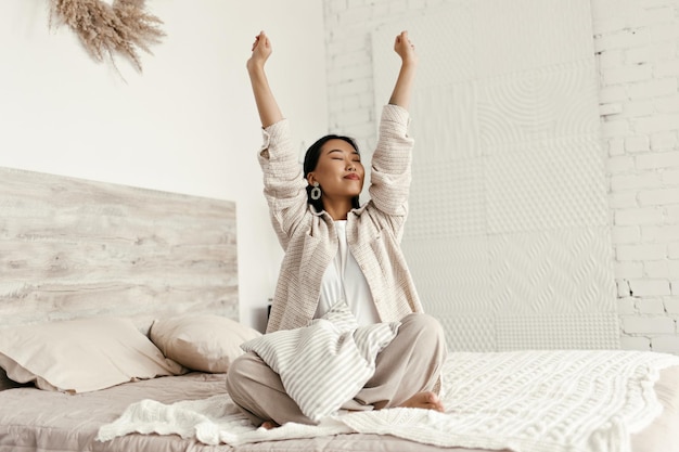 Linda mujer con chaqueta beige y pantalones cómodos se estira en el dormitorio Feliz joven asiática de buen humor se sienta en una cama suave con almohada a rayas