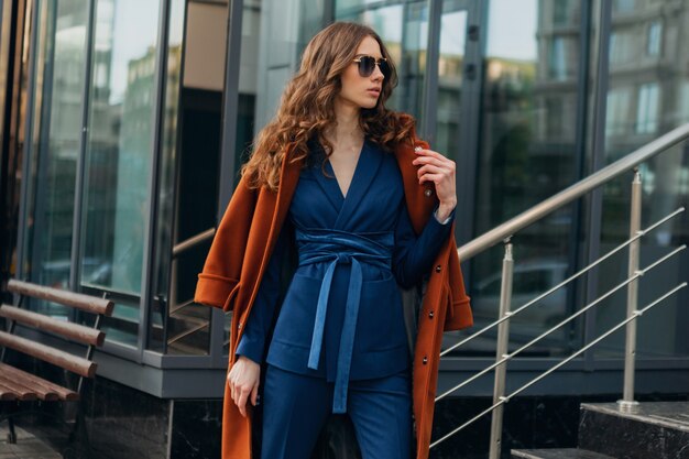 Linda mujer elegante con caminar en la calle de negocios de la ciudad urbana vestida con abrigo marrón cálido y traje azul, estilo callejero de moda primavera otoño, con gafas de sol