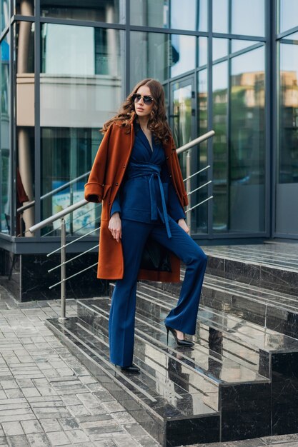 Linda mujer elegante con caminar en la calle de negocios de la ciudad urbana vestida con abrigo marrón cálido y traje azul, estilo callejero de moda primavera otoño, con gafas de sol