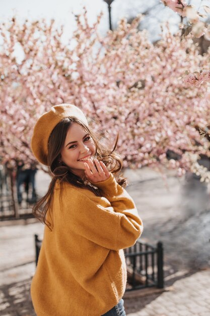 Linda mujer en elegante traje naranja y ríe en el fondo de sakura. Señora atractiva en suéter de cashemere y boina sonriendo y caminando en el parque