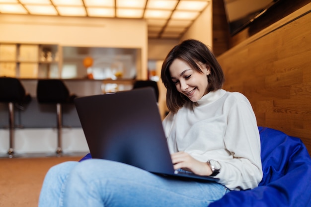 Linda mujer está sentada en una silla bolsa azul trabajando en una computadora portátil negra hablando por teléfono