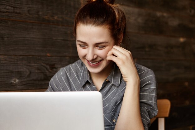 Linda mujer estudiante de jengibre con nudo de cabello mirando la pantalla de la computadora portátil genérica, sonriendo tímidamente, sus mejillas sonrojadas, con videollamada