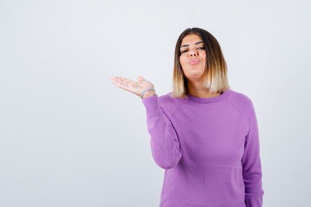 Linda mujer extendiendo la palma a un lado, haciendo pucheros con los labios en un suéter morado y luciendo pacífica, vista frontal.