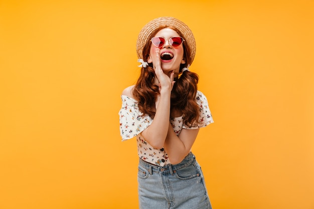 Linda mujer con gafas de sol rojas y sombrero grita. Señora vestida con falda de mezclilla, camiseta blanca y sombrero posando sobre fondo naranja.