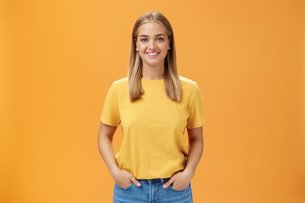 Linda mujer gordita con piel bronceada y cabello rubio posando optimista y alegre contra el fondo naranja tomados de la mano en los bolsillos sonriendo ampliamente a la cámara carismática y de aspecto amigable