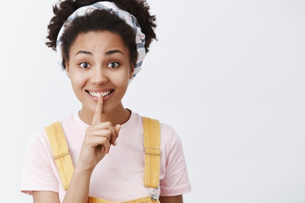 Linda mujer guapa que quiere mantener en secreto, preparando una sorpresa. Mujer de piel oscura femenina con peto amarillo y diadema, diciendo shh, haciendo un gesto de silencio, sosteniendo el dedo índice sobre la boca