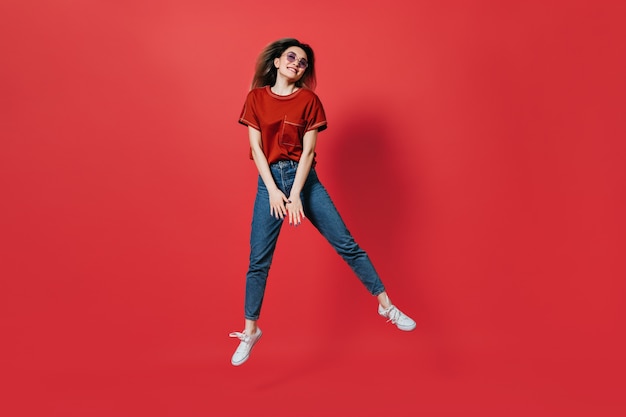 Linda mujer en jeans y camiseta brillante saltando sobre pared roja