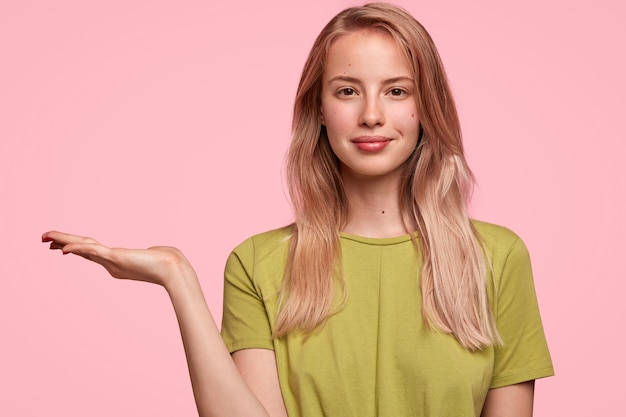 Linda mujer joven con apariencia agradable, levanta la palma, anuncia algo, vestida con una camiseta casual verde, se para contra la pared rosa