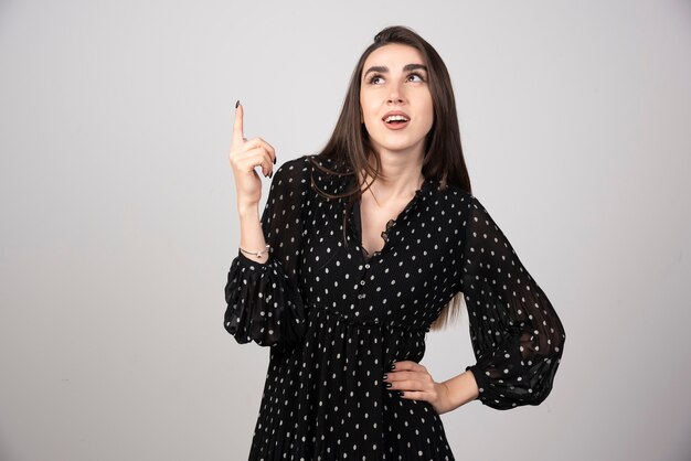 Linda mujer joven apuntando con el dedo hacia una pared gris.