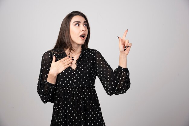 Linda mujer joven apuntando con el dedo hacia una pared gris.