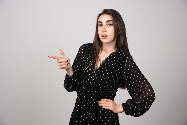 Linda mujer joven apuntando con el dedo hacia una pared gris.