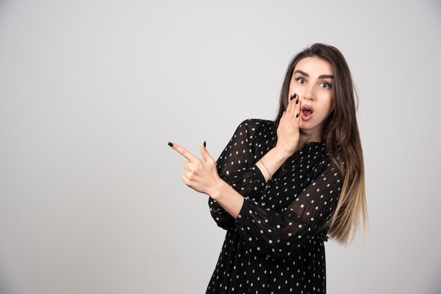 Linda mujer joven apuntando con el dedo hacia una pared gris.