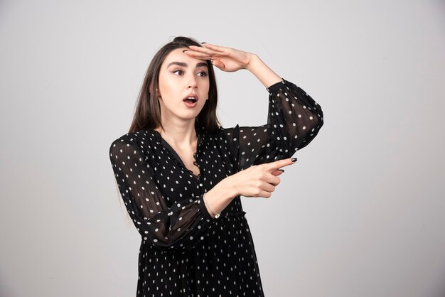 Linda mujer joven apuntando con el dedo hacia una pared gris.