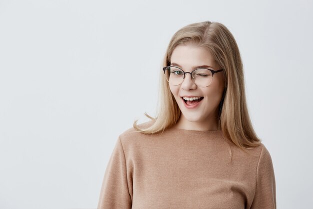 Linda mujer joven con cabello lacio rubio con suéter suelto marrón y anteojos parpadeando con placer con expresión feliz. Concepto de expresiones faciales y emociones de las personas