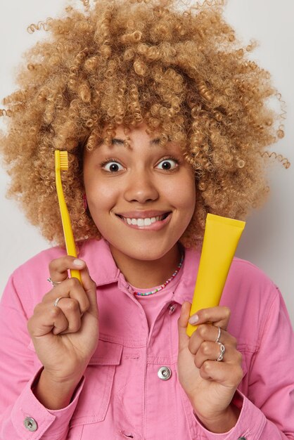 Linda mujer joven con cabello rizado y tupido sostiene pasta de dientes y cepillo de dientes amarillo que va a limpiar los dientes vestida con poses de chaqueta rosa en el interior Rutinas diarias de higiene odontología y concepto de cuidado dental