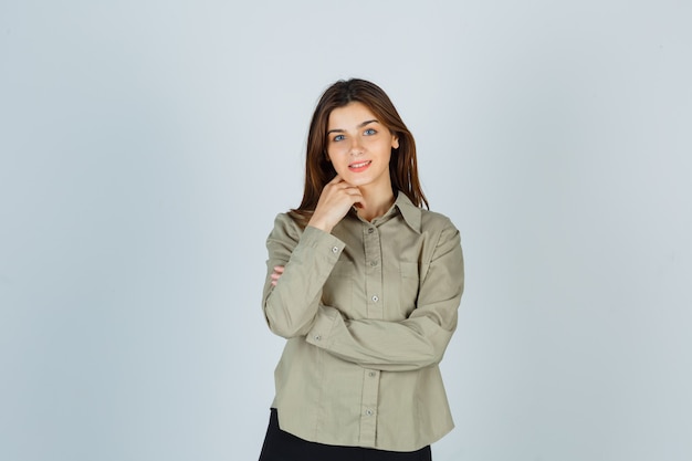 Linda mujer joven en camisa, falda de pie en pose de pensamiento y mirando alegre, vista frontal.