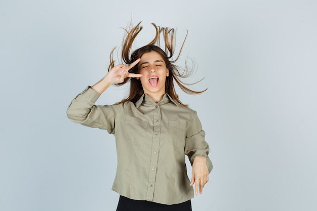 Foto gratuita linda mujer joven en camisa que muestra el signo v cerca del ojo, sacando la lengua mientras posa con el pelo al viento y luce enérgica, vista frontal.