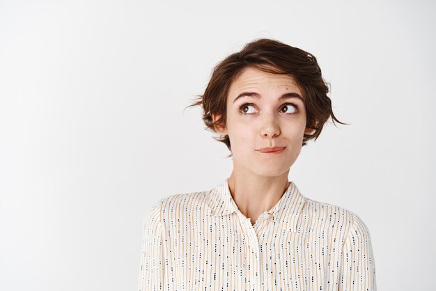 Linda mujer joven con corte de pelo de camisa que parece tentada en el espacio vacío de la esquina superior izquierda pensando en algo pensativo sobre fondo blanco