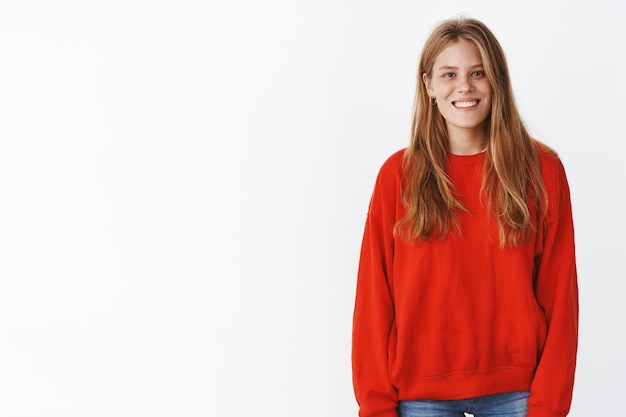 Linda mujer joven feliz de aspecto amistoso con lindas pecas y peinado largo en suéter rojo cálido sonriendo ampliamente, posando relajado y despreocupado con una expresión suave y extrovertida sobre una pared blanca