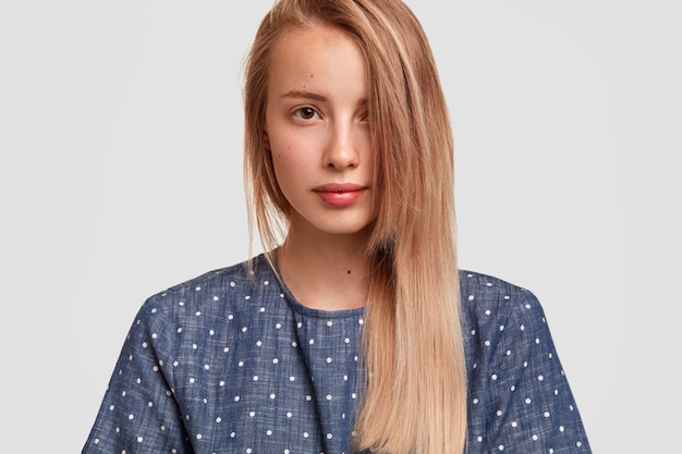 Linda mujer joven con el pelo largo peinado a un lado, mira seriamente, muestra su piel sana y perfecta, vestida con una blusa de lunares, posa contra la pared blanca. Gente, belleza, concepto de estilo de vida