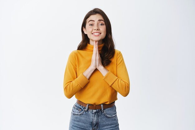 Linda mujer joven pidiendo un favor, necesita un consejo o un favor, tomados de la mano en gesto de oración y sonriendo, de pie contra la pared blanca