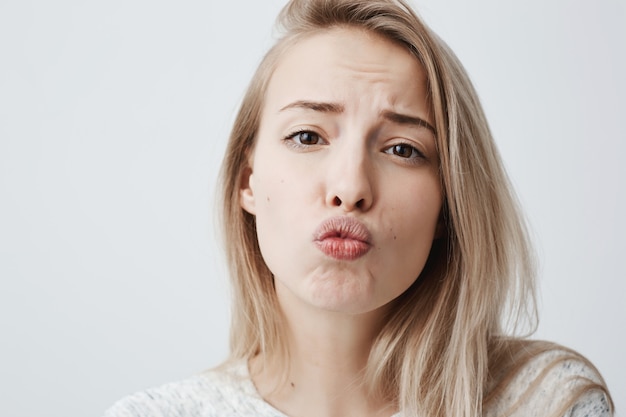 Linda mujer joven con piel pálida, cabello largo y rubio, usa un suéter casual, redondea los labios, se ve coqueta