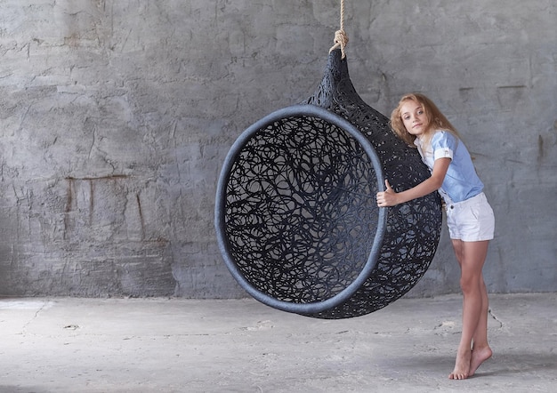 Foto gratuita linda mujer joven posando cerca de una gran silla artística negra ovalada sobre fondo gris.