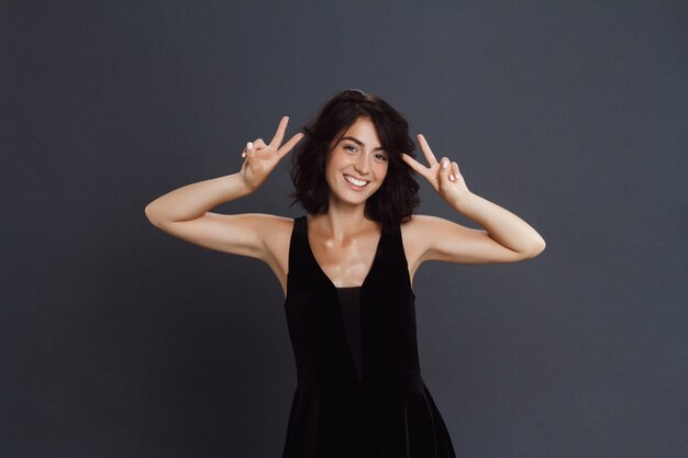 Linda mujer joven posando sobre pared gris y hacer dos gestos divertidos de paz