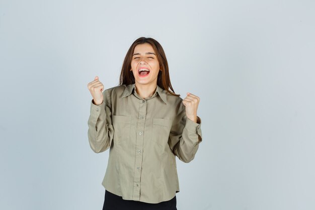 Linda mujer joven que muestra el gesto del ganador mientras grita en camisa y se ve feliz, vista frontal.