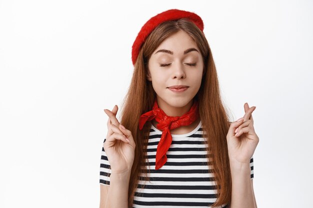 Linda mujer joven con sombrero elegante, cerrar los ojos y cruzar los dedos para la buena suerte, pedir deseos, esperar que algo suceda, orar, de pie sobre una pared blanca