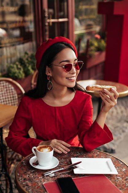 Linda mujer morena bronceada en elegante vestido rojo, boina y gafas de sol se sienta en la cafetería