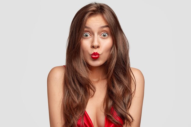 Linda mujer morena sorprendida mantiene los labios rojos redondeados, tiene los ojos abiertos, la piel pura, ve algo increíble en el frente, posa para la revista de moda, aislado sobre una pared blanca. Concepto de belleza