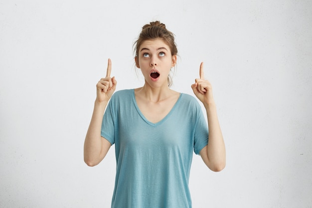 Linda mujer de ojos azules con nudo de pelo vistiendo camiseta azul casual mirando hacia arriba con asombro levantando sus dedos índices apuntando a algo habiendo abierto la boca