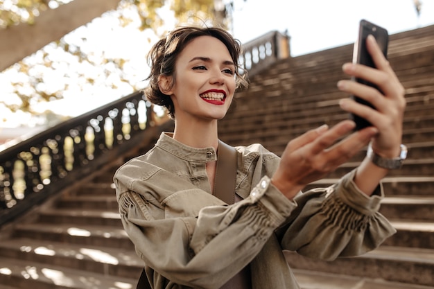 Linda mujer con peinado corto y labios rojos sonriendo sinceramente al aire libre. Mujer fresca en chaqueta vaquera haciendo selfie