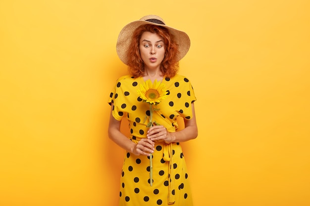 Linda mujer pelirroja sorprendida posando en vestido de lunares amarillo y sombrero de paja