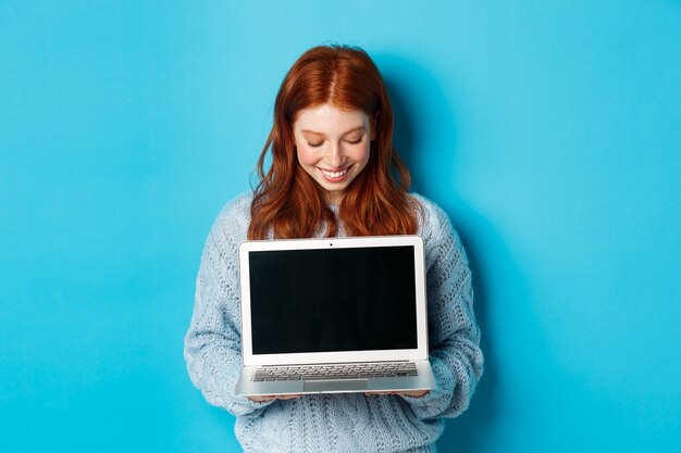 Linda mujer pelirroja en suéter, mostrando y mirando la pantalla del portátil con una sonrisa complacida, demostrando algo en línea, de pie sobre fondo azul.