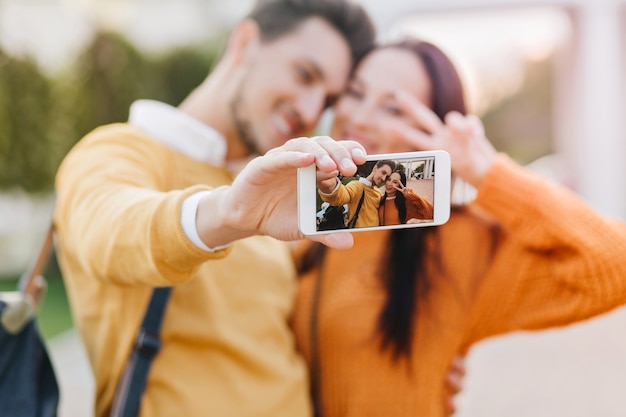 Linda mujer posando con el signo de la paz mientras su novio en suéter naranja haciendo selfie