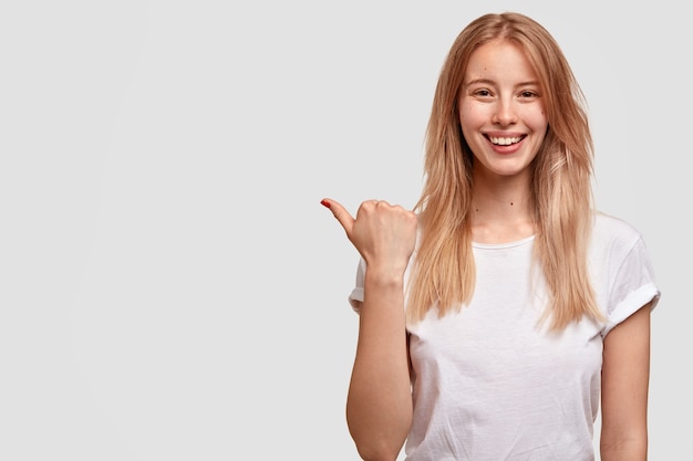 Linda mujer positiva con cabello claro, señala a un lado con el pulgar, vestida con ropa casual, tiene expresión encantada