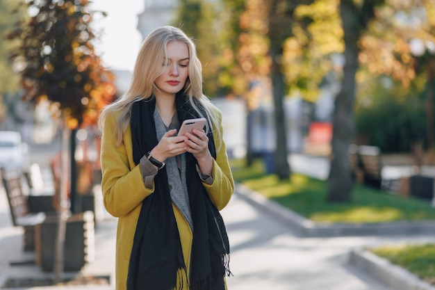 Linda mujer rubia atractiva emocional en abrigo con smartphone camina por la calle de la ciudad