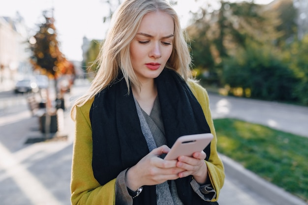 Linda mujer rubia atractiva emocional en abrigo con smartphone camina por la calle de la ciudad