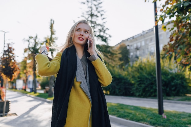 Linda mujer rubia atractiva emocional en abrigo con smartphone camina por la calle de la ciudad