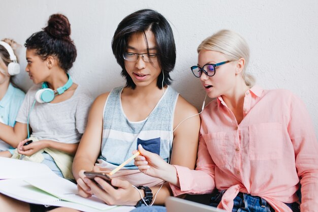 Linda mujer rubia con gafas apuntando con un lápiz a la pantalla del teléfono mientras escucha música con un chico asiático. Estudiantes alegres que aprenden juntos y se divierten en la universidad.