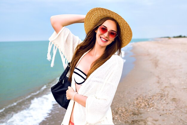 Linda mujer rubia haciendo selfie en la playa del océano, vestida con traje boho y gafas de sol divertidas, sombrero de paja vintage, enviando un beso.
