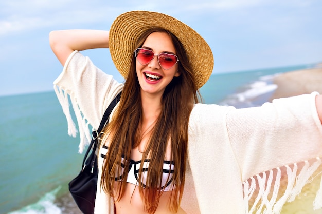 Linda mujer rubia haciendo selfie en la playa del océano, vestida con traje boho y gafas de sol divertidas, sombrero de paja vintage, enviando un beso.