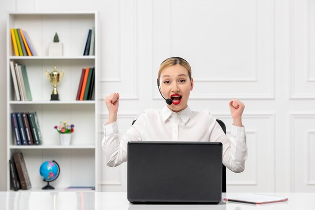 Linda mujer de servicio al cliente con camisa blanca con auriculares y computadora emocionada sosteniendo los puños