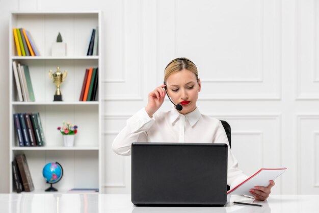 Linda mujer de servicio al cliente en camisa blanca con auriculares y computadora leyendo notas y escuchando