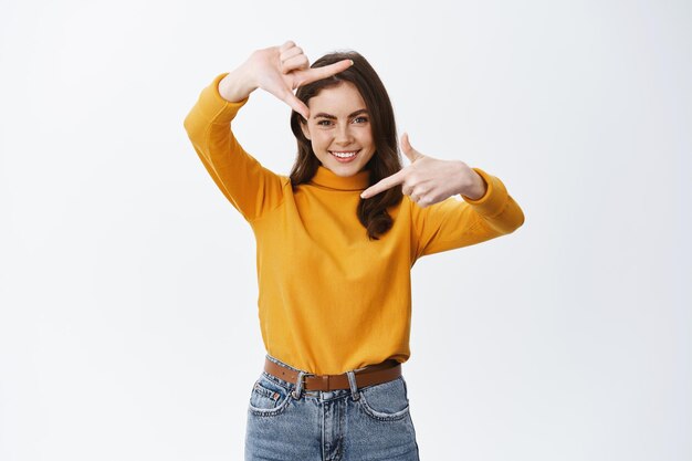 Linda mujer sonriente mostrando la mano enmarca la cámara, tomando una foto del momento, buscando la perspectiva, de pie en blanco