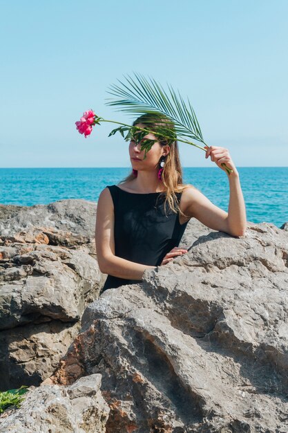 Linda mujer sosteniendo flores y hojas de palma, apoyándose en la roca cerca del mar