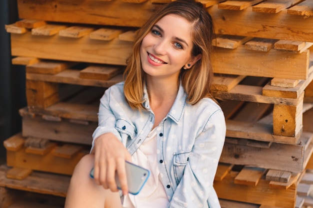 Linda mujer sosteniendo el teléfono en las manos mientras está sentado en paletas de madera y sonriendo a la cámara