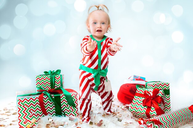 Linda niña de 1 año con sombrero de santa posando sobre fondo de Navidad. De pie en el suelo con bola de Navidad. Temporada de vacaciones.